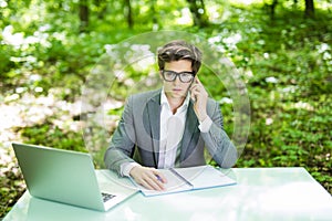 Portrait of young handsome manager working at laptop at office table and talk at phone with costumer in green forest park. Busines