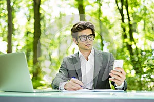 Portrait of young handsome manager working at laptop at office table and talk at phone with costumer and make notice in green fore
