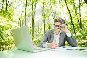 Portrait of young handsome sad business man in suit working at laptop at office table in green forest park. Business concept.