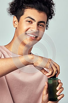 Portrait of young handsome smiling guy with a bottle of facial lotion in his hands. Swarthy man with black curly hair