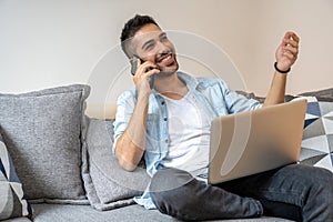 Portrait of young, handsome, smiling man talking on the phone and using his laptop while sitting on the sofa at home.