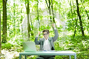 Portrait of young handsome succesful business man in suit working at laptop at office table with raised hands in green forest park