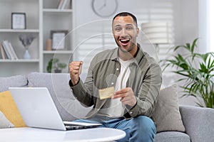 Portrait of young happy African American man sitting on couch at home in front of laptop, holding credit card and