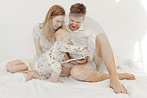 Portrait of young happy attractive family with cute cherubic infant baby toddler in white clothes sitting on white bed.