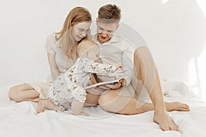 Portrait of young happy attractive family with cute cherubic infant baby toddler in white clothes sitting on white bed. photo