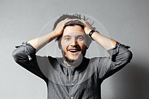 Portrait of young happy bearded guy with disheveled hair. Hold head with hands. Grey background.