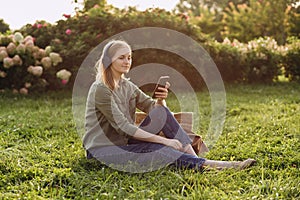 Portrait of young happy blonde woman listening to music with headphones and smiling on a green grass outdoor. Music