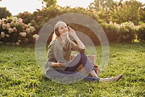 Portrait of young happy blonde woman listening to music with headphones and smiling on a green grass outdoor. Music