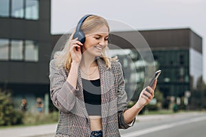 Portrait of young happy blonde woman listening to music with headphones and smiling while walking on the street in the