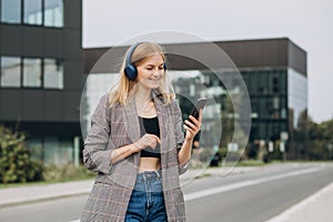 Portrait of young happy blonde woman listening to music with headphones and smiling while walking on the street in the