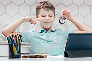 Portrait of young happy boy with closed eyes sitting at desk with digital tablet, pointing index finger at small clock.