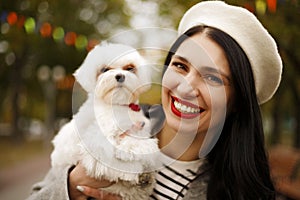 Portrait of young happy Caucasian woman in french white hat and warm coat holding and hugging her little cute white dog maltese