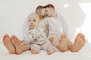 Portrait of young happy couple looking at each other in white clothes with cherubic baby infant toddler sitting on bed.