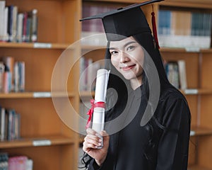 Portrait of a young happy and excited Asian woman university graduates in graduation gown and cap holds and shows a degree