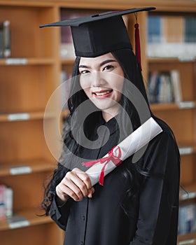 Portrait of a young happy and excited Asian woman university graduates in graduation gown and cap holds and shows a degree
