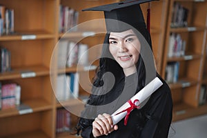 Portrait of a young happy and excited Asian woman university graduates in graduation gown and cap holds and shows a degree