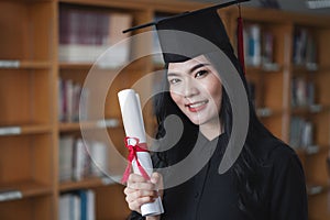 Portrait of a young happy and excited Asian woman university graduates in graduation gown and cap holds and shows a degree