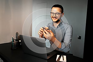 Portrait of young happy man working home at laptop, using smartphone, a message wearing eyeglasses and shirt.