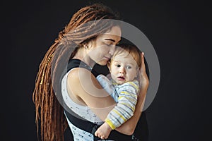 Portrait young happy mom with baby son in ergo backpack on dark wall on the background. Concept of modern parents