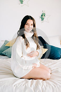 Portrait of a young happy pregnant woman in a white warm sweater sitting on the bed and holding her stomach