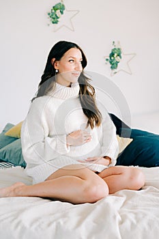 Portrait of a young happy pregnant woman in a white warm sweater sitting on the bed and holding her stomach