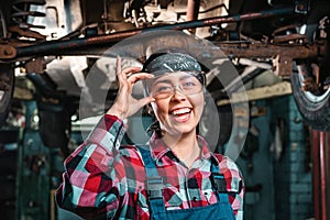 Portrait of young happy repairwoman, in a uniform and glasses poses standing under a car on a lift. Indoors garage