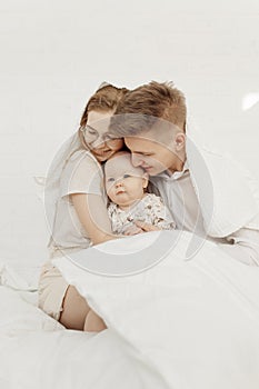 Portrait of young happy smiling beautiful family with cute cherubic infant baby in white clothes sitting on white bed.