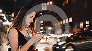 Portrait of young happy woman standing in traffic downtown in the evening in New York, America and using the smartphone.