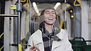 Portrait of young happy woman wearing in coat with headphones listening to music and funny dancing in public transport.