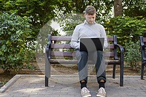 Portrait of a young hipster freelancer working on his laptop sitting on a bench in a park