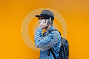 Portrait of young hipster talking on smartphone on orange background. Wearing jeans jacket, cap, backpack and glasses.