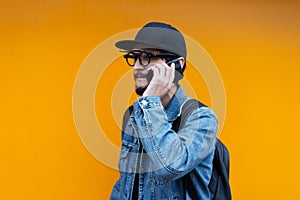Portrait of young hipster talking on smartphone on orange background. Wearing jeans jacket, cap, backpack and glasses.