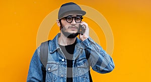 Portrait of young hipster talking on smartphone on orange background. Wearing jeans jacket, cap, backpack and glasses.