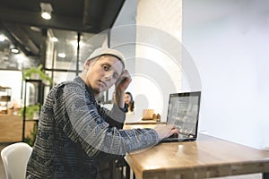 Portrait of a young hipster who works for a laptop in a cozy cafe and looks at the camera. Freelancer at work in a cafe.