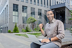 Portrait of a young hispanic male student sitting with a laptop on a bench outside the campus, looking forward to the