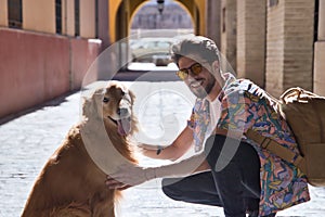Portrait of young Hispanic man with beard and sunglasses crouching with his dog looking at camera on a sunny street at sunset.