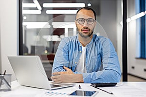 Portrait of young hispanic man in glasses and shirt, office worker and student sitting at desk with laptop looking