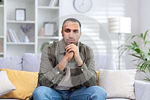 Portrait of a young hispanic man looking seriously at the camera, sitting at home on the couch with folded hands