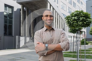Portrait of young hispanic student man smiling and looking at camera near university campus with arms crossed wearing