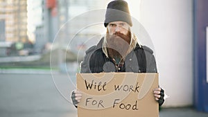 Portrait of young homeless man with cardboard looking at camera and wants to work for food looking at camera at street