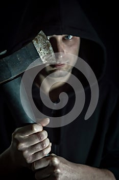 Portrait of a young hooded man with blue eyes who with an ax in his hand has a disturbing look - focus on the blade