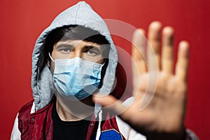 Portrait of young hooded man wearing medical flu mask, showing STOP gesture with hand on background of red color.