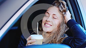 Portrait of a young independent woman in the car. Sits at the wheel, looks at the camera, holds a glass of coffee in his