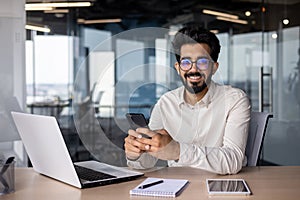 Portrait of a young Indian businessman, office worker sitting at a desk and talking on the phone, smiling and looking at