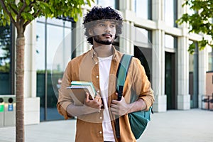 Portrait of a young Indian male student standing outside the campus, looking seriously to the side, holding books and a