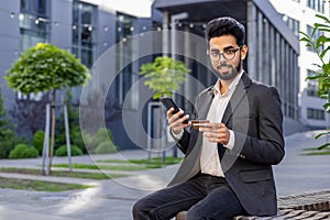 Portrait of a young Indian man in a business suit sitting on a bench near an office center, holding a credit card and a