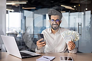 Portrait of a young Indian man sitting in the office at the table, holding cash money and a phone in his hands, smiling