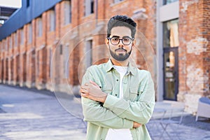 A portrait of a young Indian man standing on a city street, crossing his arms and looking confidently into the camera