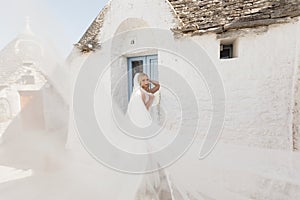Portrait of young joyful woman bride with flying bridal veil, standing near old white stone house trullo in sunny Italy.