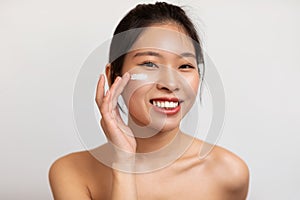 Portrait of young korean lady applying facial cream under her eyes, taking care of skin, standing over white background